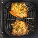 Two cooked bone-in pork chops seasoned with herbs and spices sit in an air fryer basket.