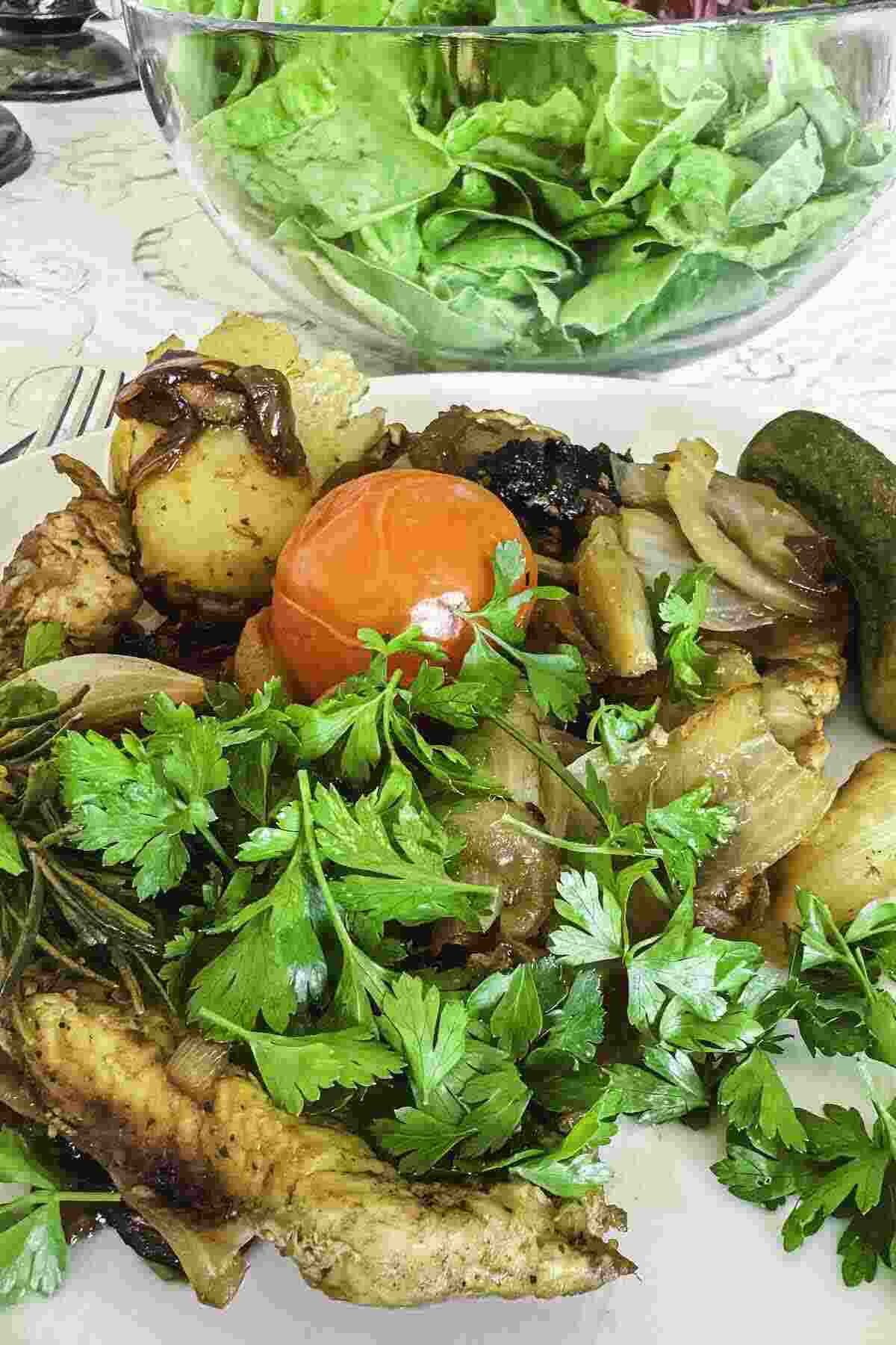 A plate of roasted chicken, potatoes, onions, fresh parsley, and a tomato, with a side of pickles and a bowl of green lettuce salad in the background.