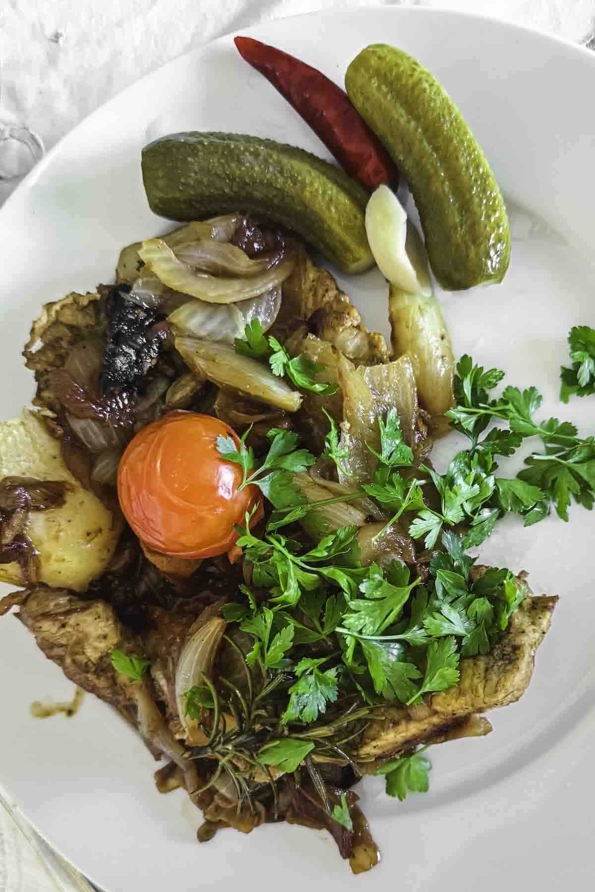 A plate with roasted vegetables including onions and tomato, garnished with parsley, served with pickles and a red chili pepper.