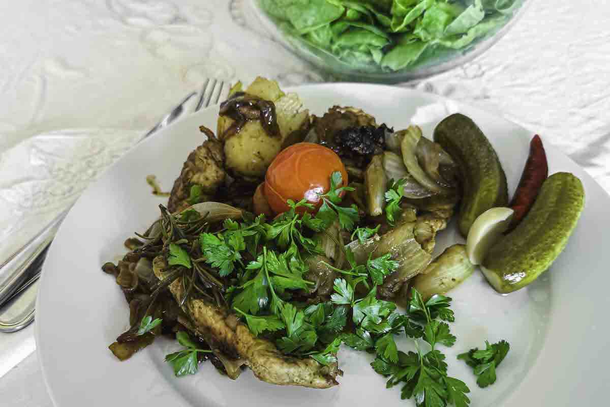 A plate with cooked meat, onions, parsley, a cherry tomato, pickles, garlic, and a red chili, with a bowl of leafy greens in the background.