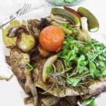 A plate of cooked meat with onions, a whole tomato, fresh herbs, pickles, whole chili peppers, and potatoes, garnished with rosemary.