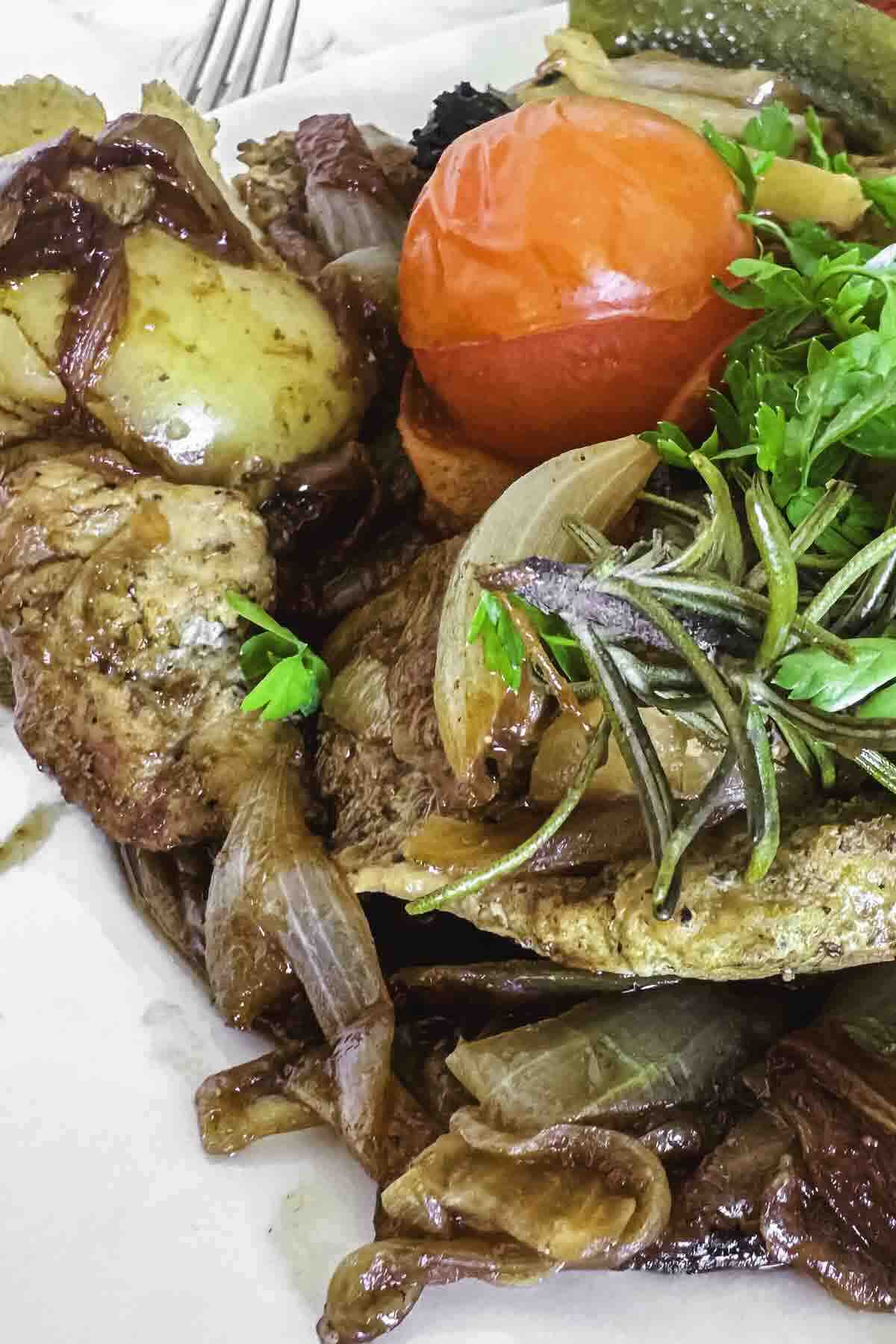 Cooked meat with caramelized onions, a whole roasted tomato, herbs, and a side of vegetables served on a white plate.