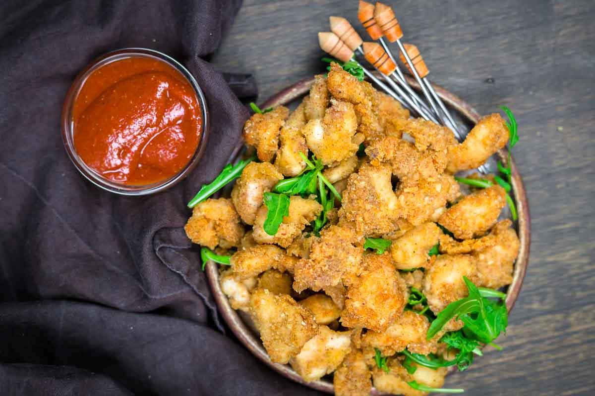 A plate of breaded, fried chicken pieces garnished with greens, served with skewers and a small bowl of red dipping sauce on the side.