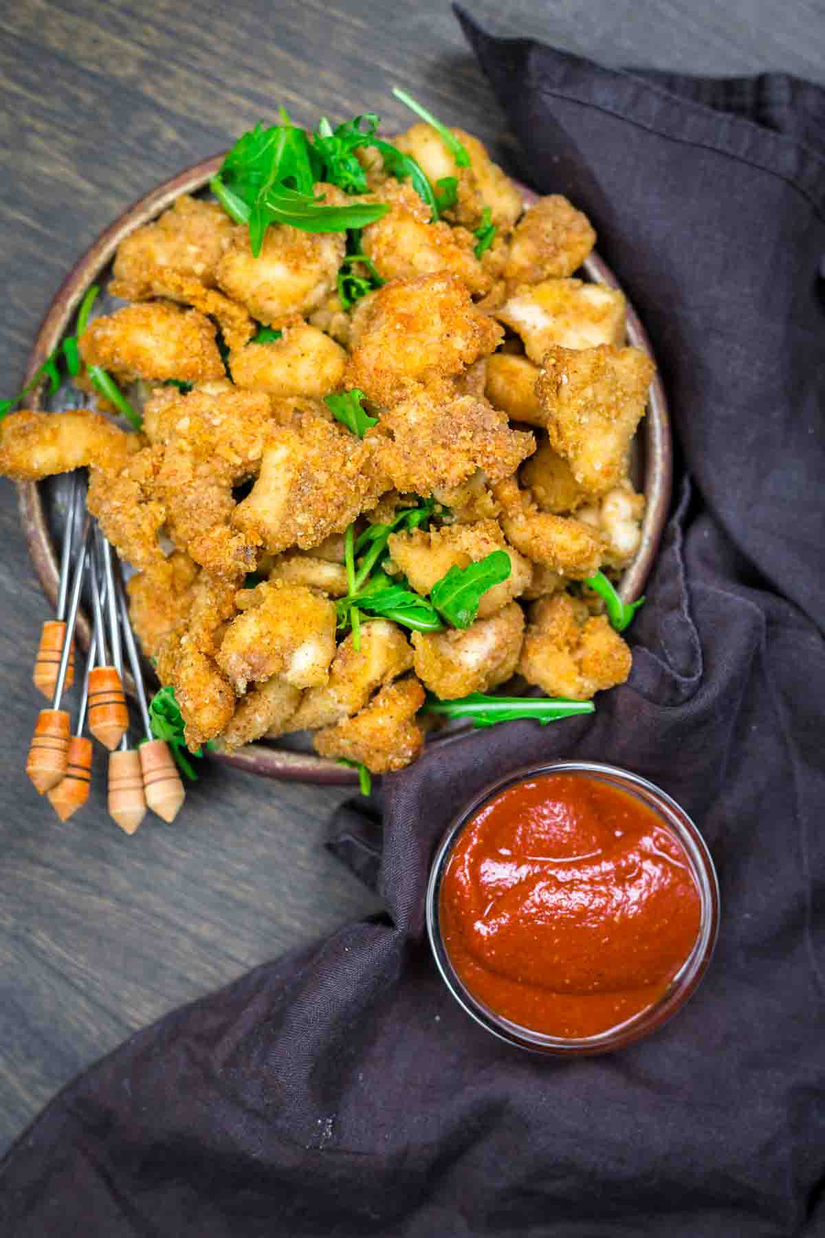 A plate of breaded, fried chicken bites garnished with greens, served with skewers and a small bowl of red dipping sauce on a dark cloth.