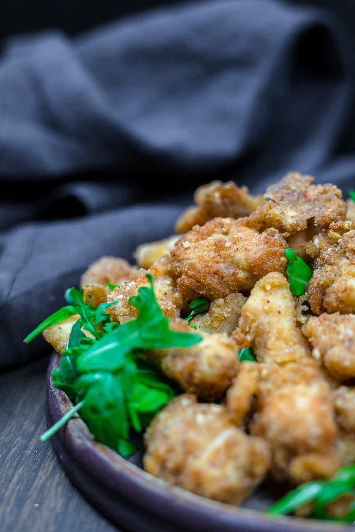 A close-up of a plate with golden-brown fried chicken pieces and fresh green arugula, set on a dark wooden surface with a dark cloth in the background.