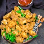 A plate of breaded and fried chicken bites garnished with fresh greens, served with dipping sauce and skewers on the side.