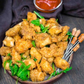 A plate of breaded and fried chicken bites garnished with fresh greens, served with dipping sauce and skewers on the side.