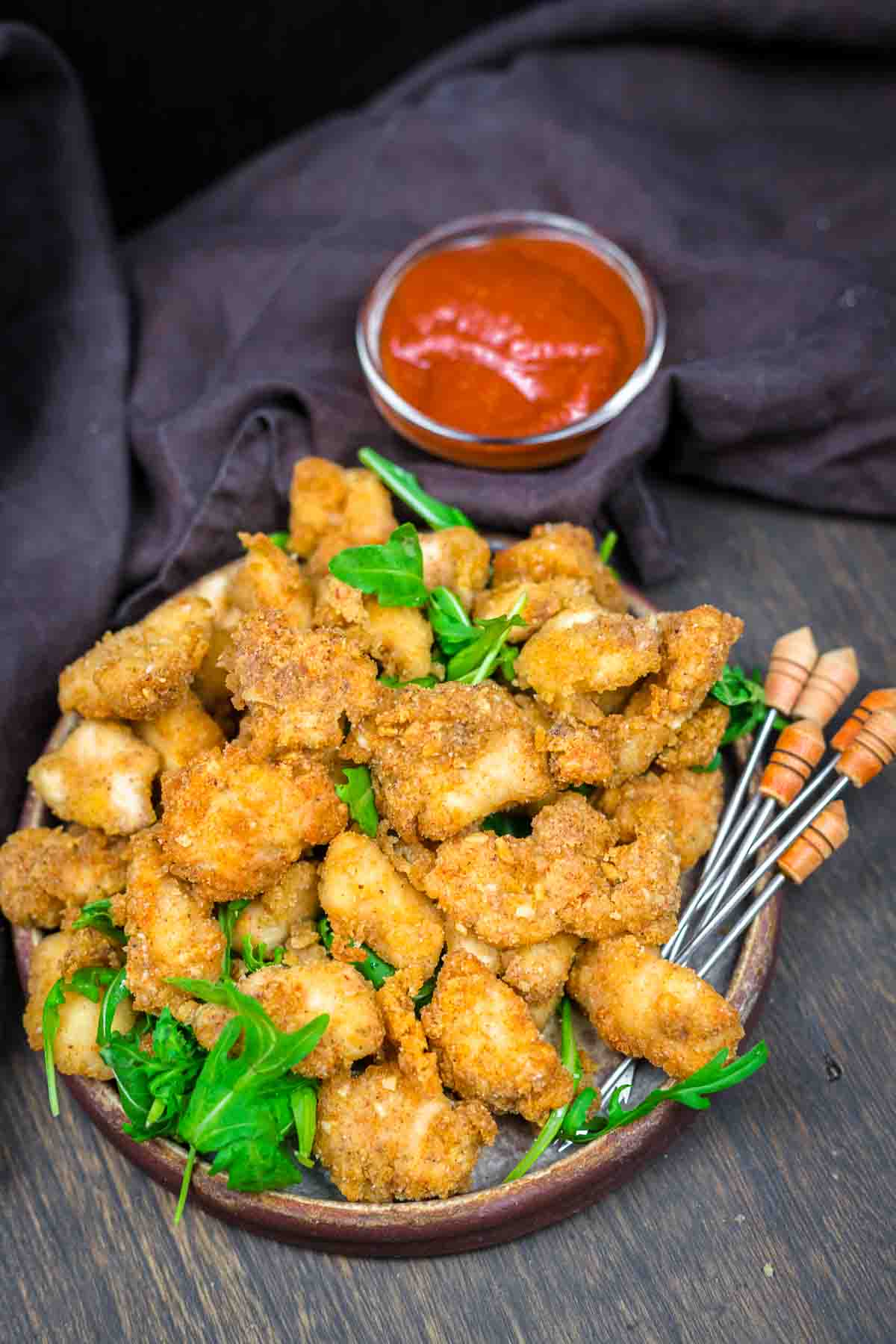 A plate of breaded and fried chicken bites served with green garnish and a bowl of red dipping sauce on the side, with skewers placed nearby.