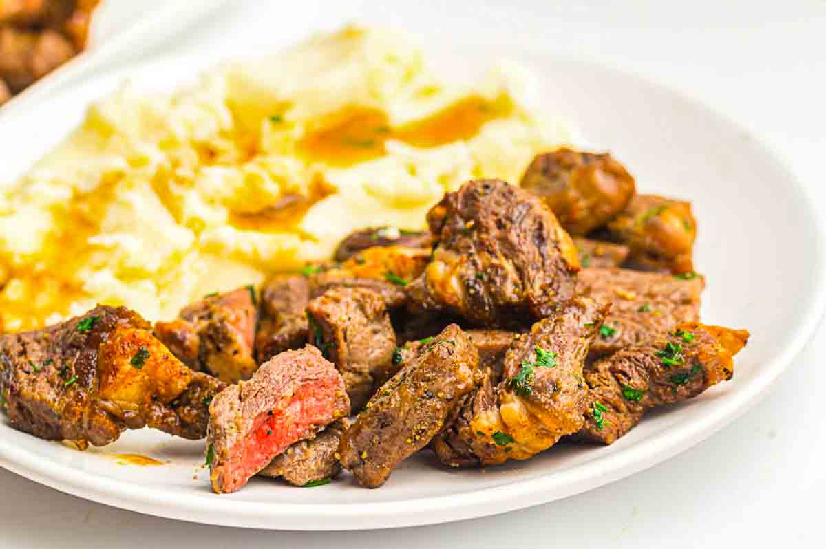 A white plate with grilled steak bites and a serving of mashed potatoes topped with brown gravy.