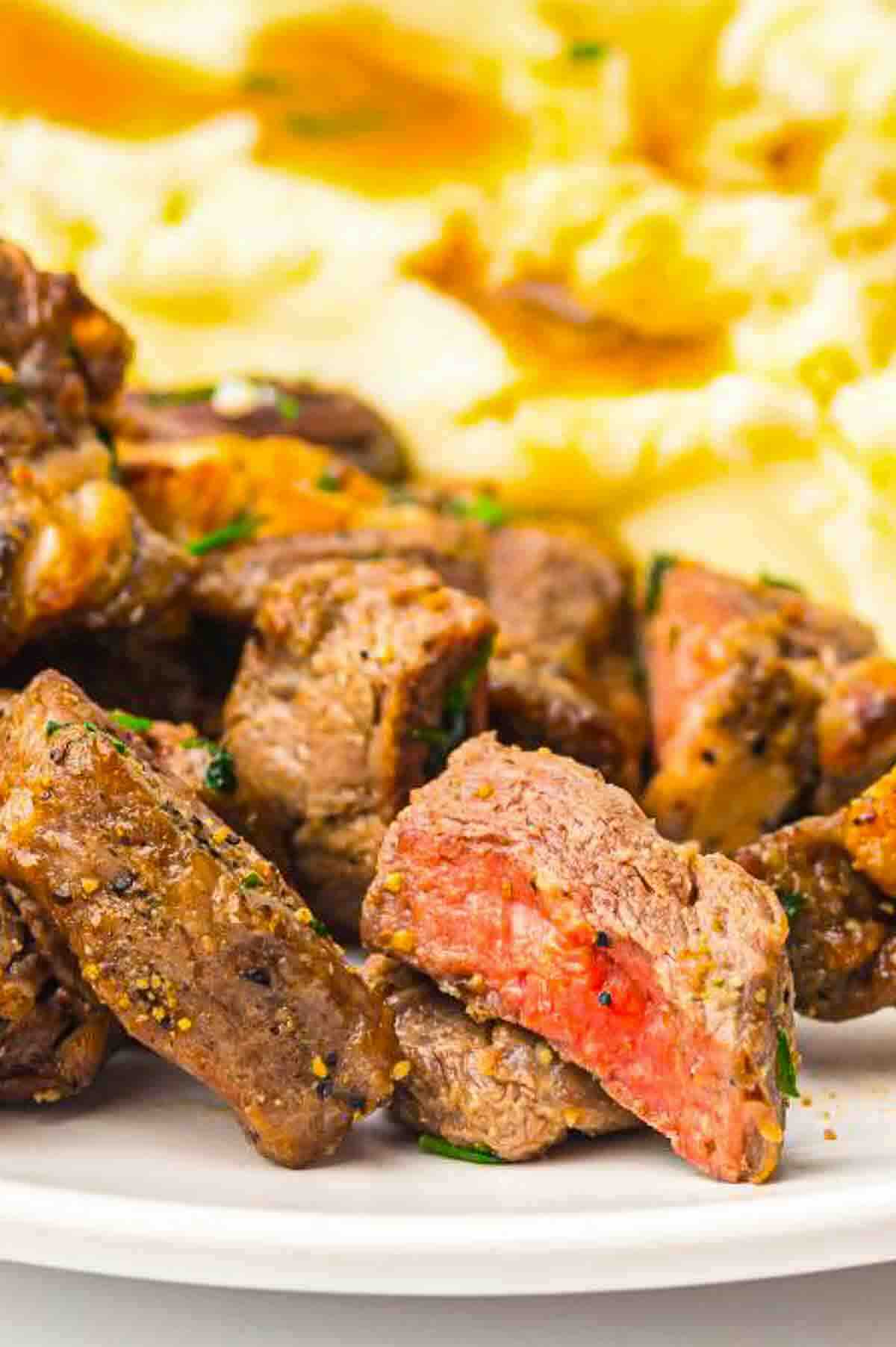 Close-up of cooked steak bites with a pink center on a white plate, served alongside mashed potatoes with brown gravy in the background.