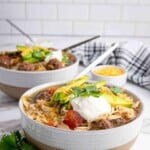 Two bowls of shredded chicken and beef soup topped with avocado, sour cream, cheese, and cilantro, with spoons, on a marble counter near a striped towel and a small bowl of cheese.