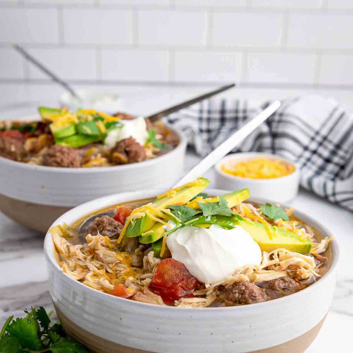 Two bowls of shredded chicken and beef soup topped with avocado, sour cream, cheese, and cilantro, with spoons, on a marble counter near a striped towel and a small bowl of cheese.