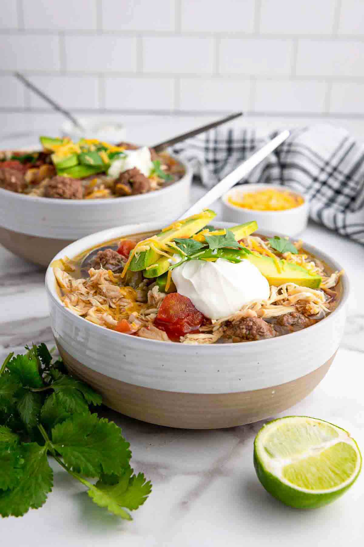 A bowl of soup topped with shredded cheese, sliced avocado, sour cream, and cilantro, with a spoon inside. A halved lime, fresh cilantro, and another bowl are in the background.