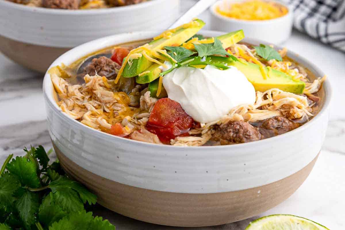 A bowl of chicken soup topped with sliced avocado, shredded cheese, sour cream, chopped parsley, and meatballs, with cilantro and lime nearby.