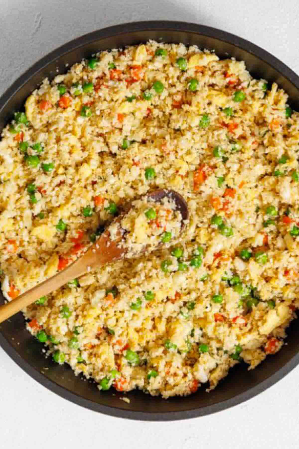 A pan filled with cauliflower fried rice mixed with peas, carrots, and scrambled eggs, with a wooden spoon resting inside.