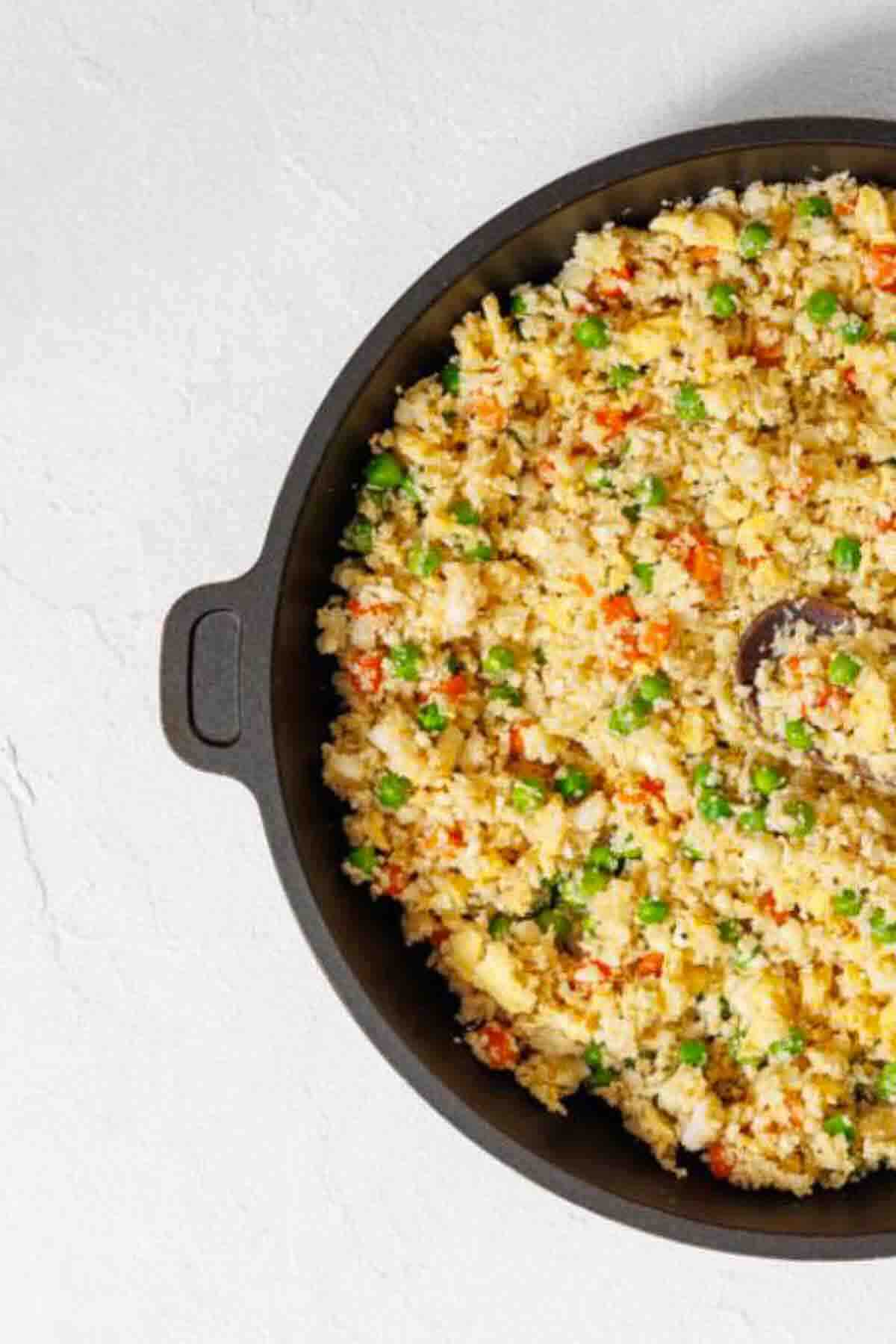 A black skillet filled with vegetable fried rice, including peas, carrots, and scrambled eggs, with a wooden spoon partially visible.