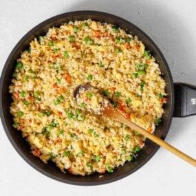 A large pan filled with cauliflower fried rice mixed with peas, carrots, and scrambled eggs, with a wooden spoon resting inside.