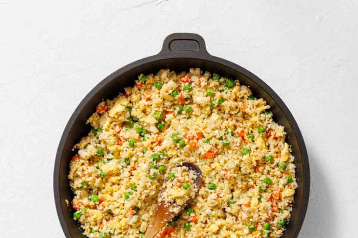 A skillet filled with cauliflower fried rice mixed with peas, carrots, and scrambled eggs, with a wooden spoon resting inside.