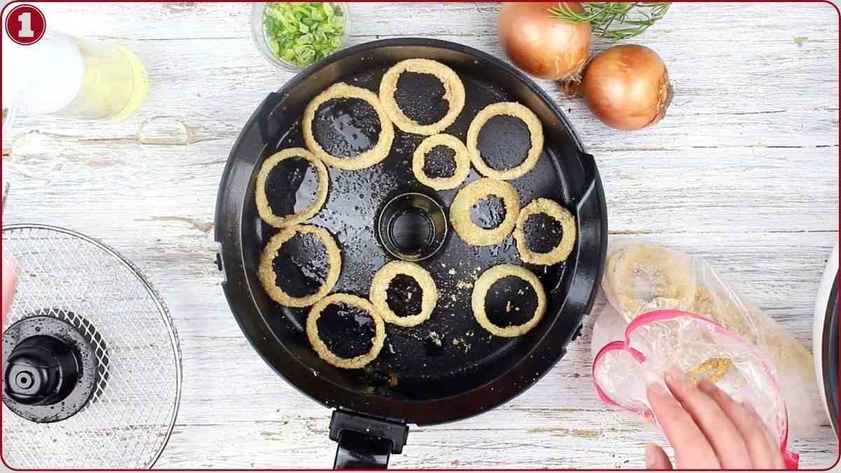 A top-down view of a round air fryer basket with breaded onion rings inside, surrounded by onions, chopped green onions, oil, and kitchen utensils on a light wooden surface.