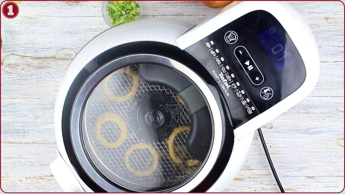 Top view of an air fryer with several onion rings inside on a mesh tray, placed on a light-colored wooden surface with chopped green onions nearby.