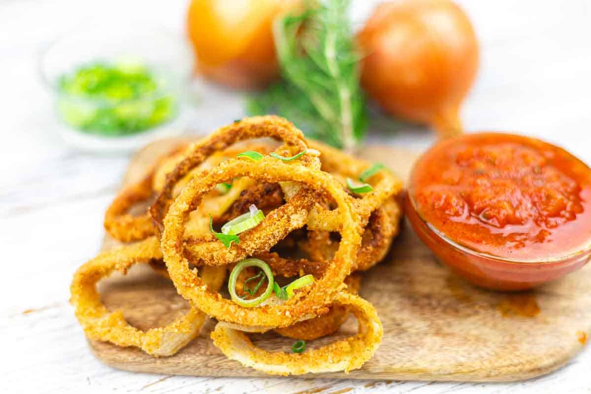 A stack of crispy fried onion rings garnished with green onions on a wooden board, served with a bowl of red dipping sauce.