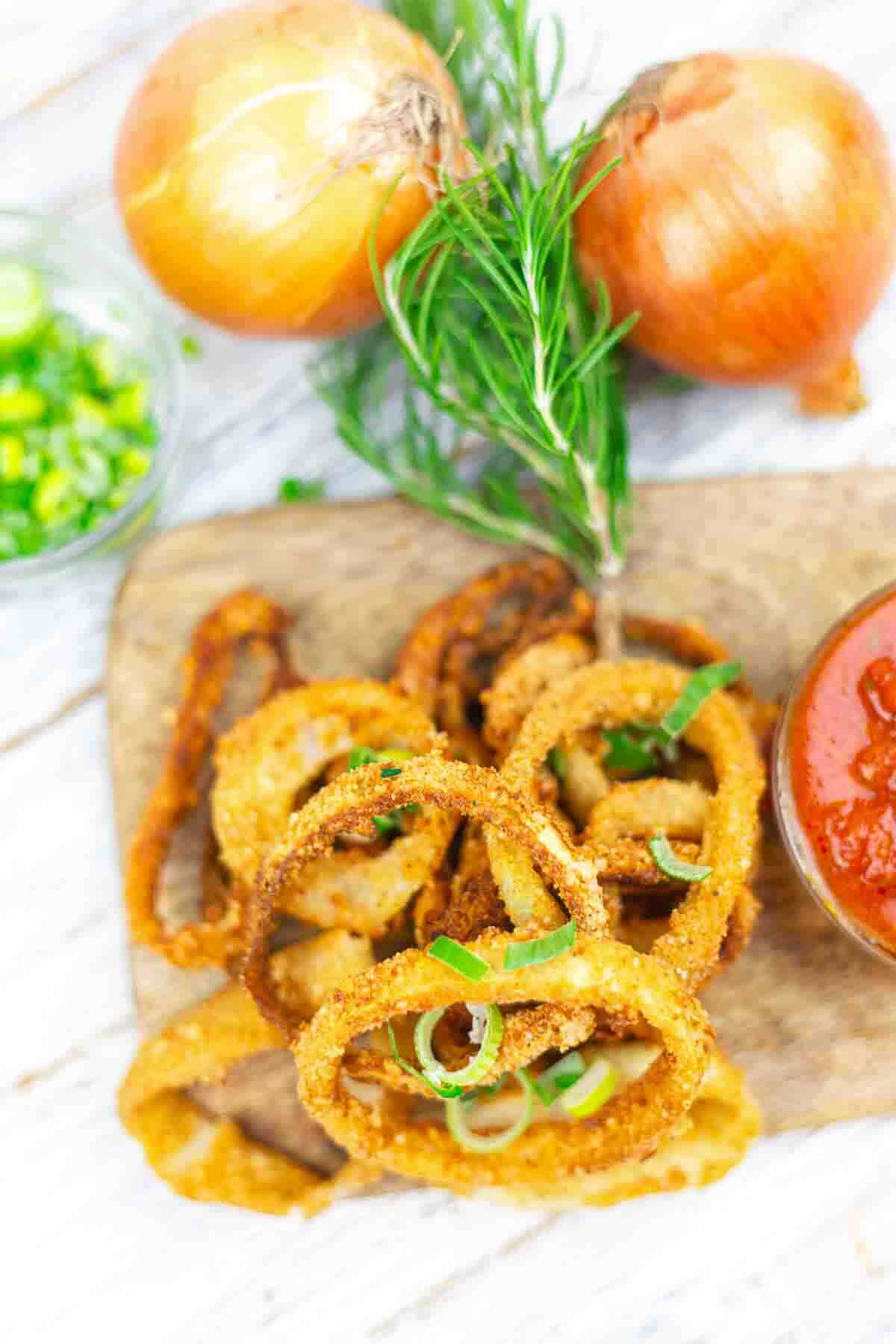A serving of crispy fried onion rings garnished with green onions, next to a bowl of red dipping sauce, fresh onions, and rosemary on a wooden board.