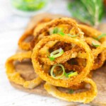A pile of crispy, golden-brown onion rings garnished with chopped green onions on a wooden board.