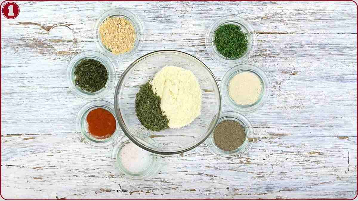 Eight small bowls containing various dried herbs, spices, and powders are arranged in a circle on a white wooden surface, with one larger bowl in the center.