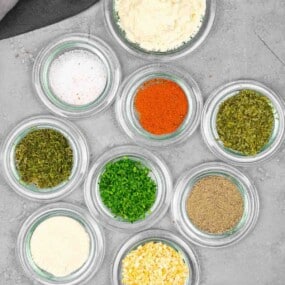 Nine small glass bowls filled with various spices and dried herbs, including salt, paprika, parsley, black pepper, and others, arranged on a gray surface.