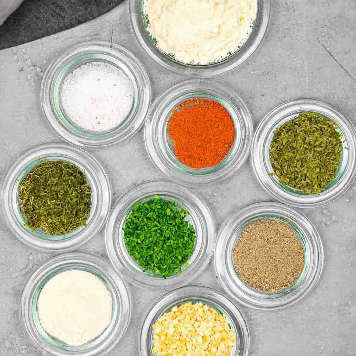 Nine small glass bowls filled with various spices and dried herbs, including salt, paprika, parsley, black pepper, and others, arranged on a gray surface.