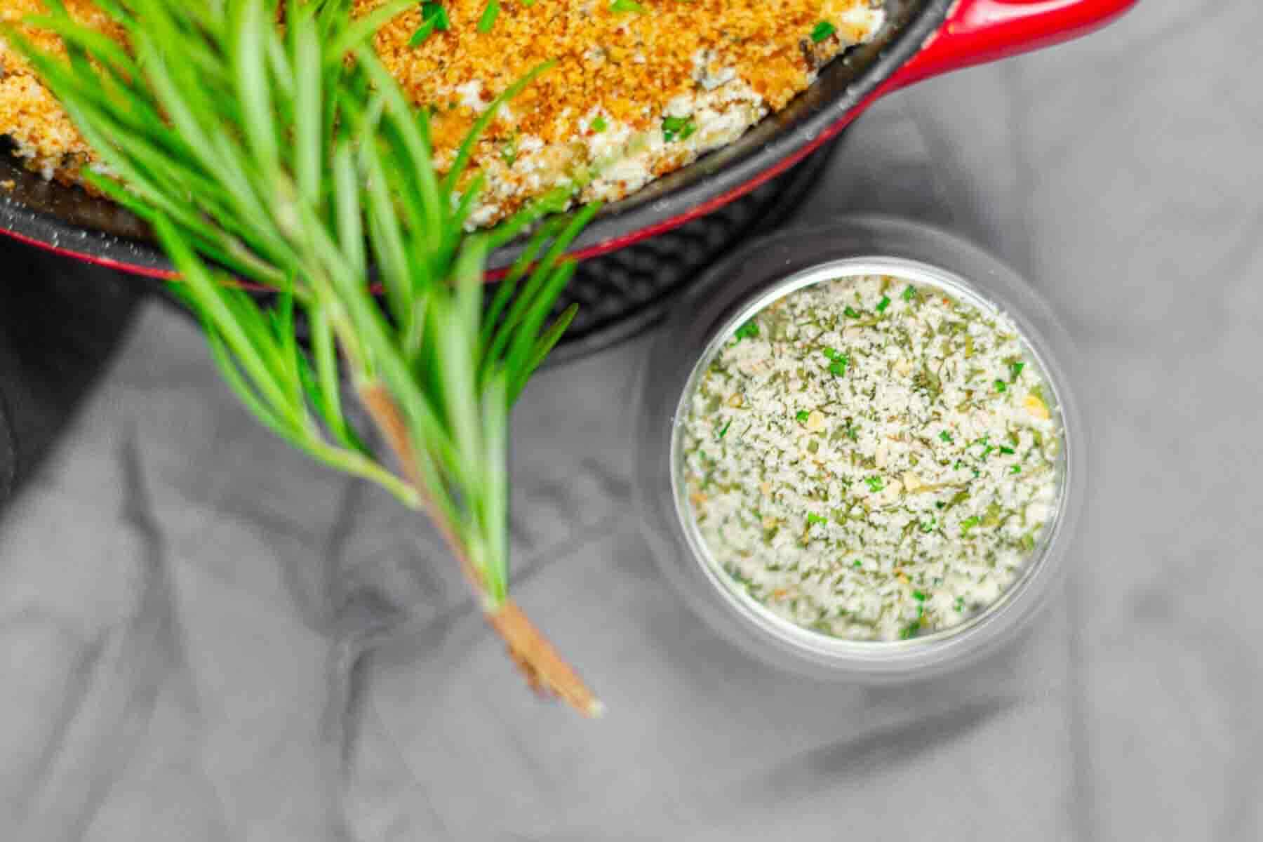 A sprig of rosemary lies beside a bowl of herbed breadcrumb mixture and a red baking dish filled with a baked casserole topped with breadcrumbs.