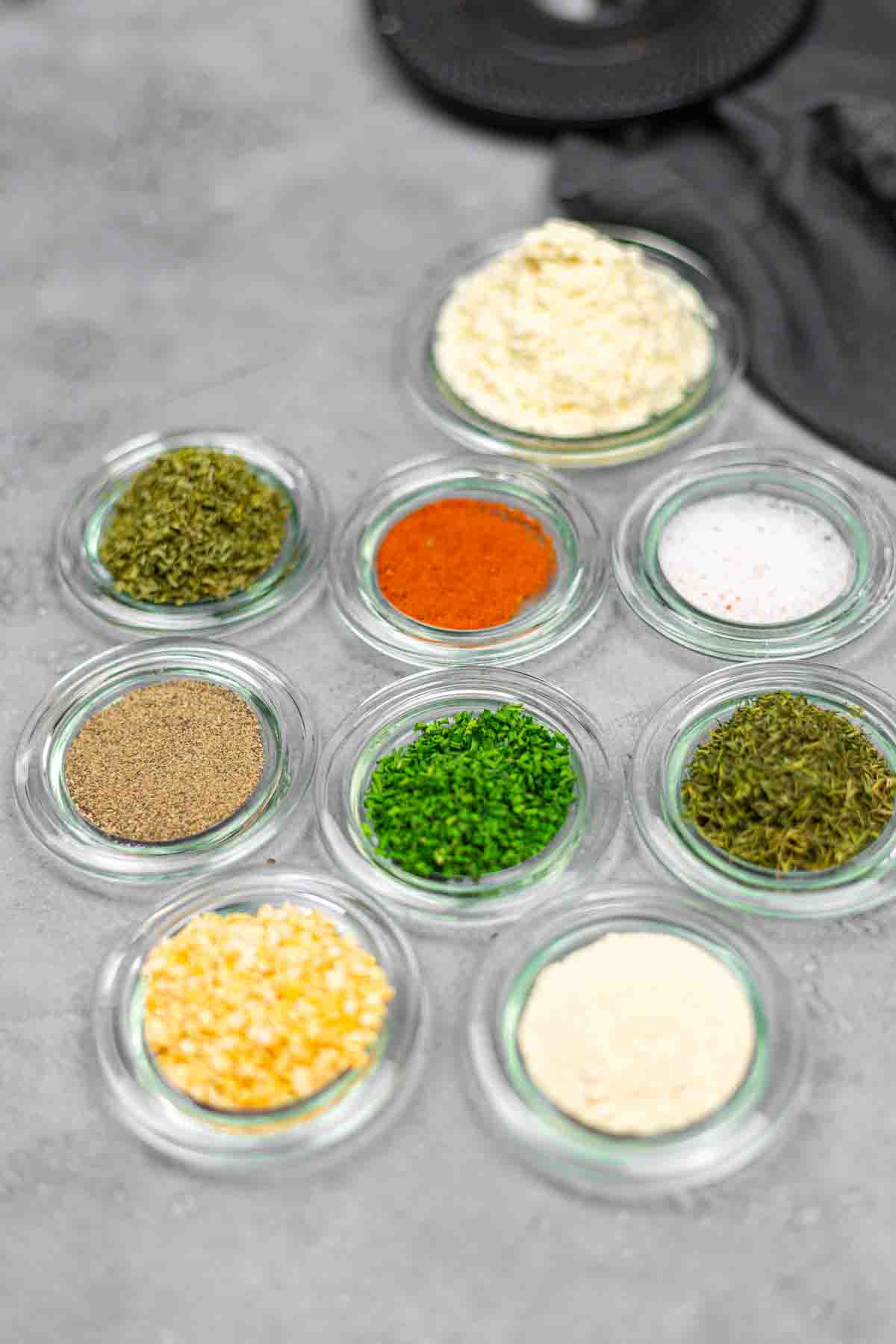 Nine small glass bowls with various dried herbs, spices, and seasonings arranged on a gray surface.