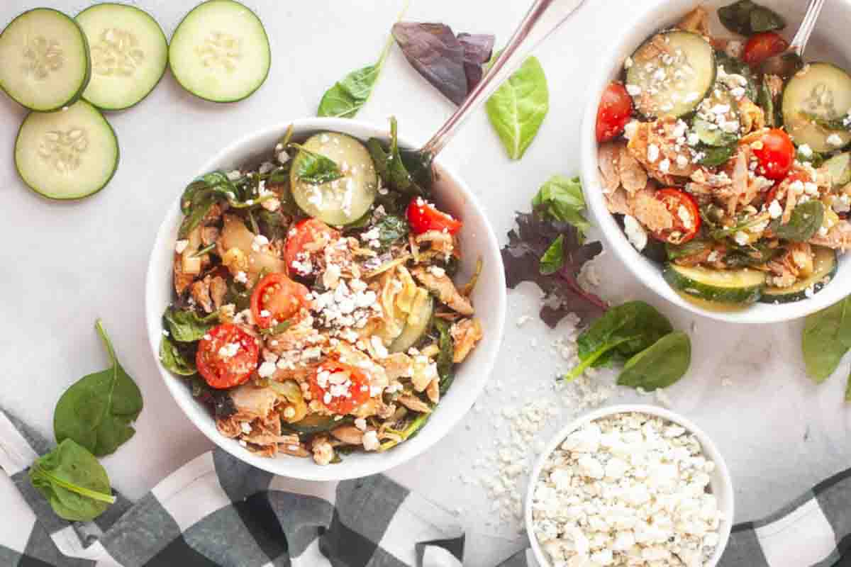 Two bowls of salad with shredded chicken, cherry tomatoes, cucumber slices, leafy greens, and crumbled cheese on a white surface, surrounded by loose greens and sliced cucumbers.