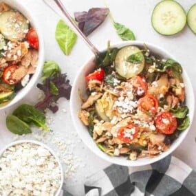 A bowl of salad with cherry tomatoes, cucumber slices, leafy greens, shredded chicken, and crumbled feta cheese, with extra feta and salad leaves on the table.