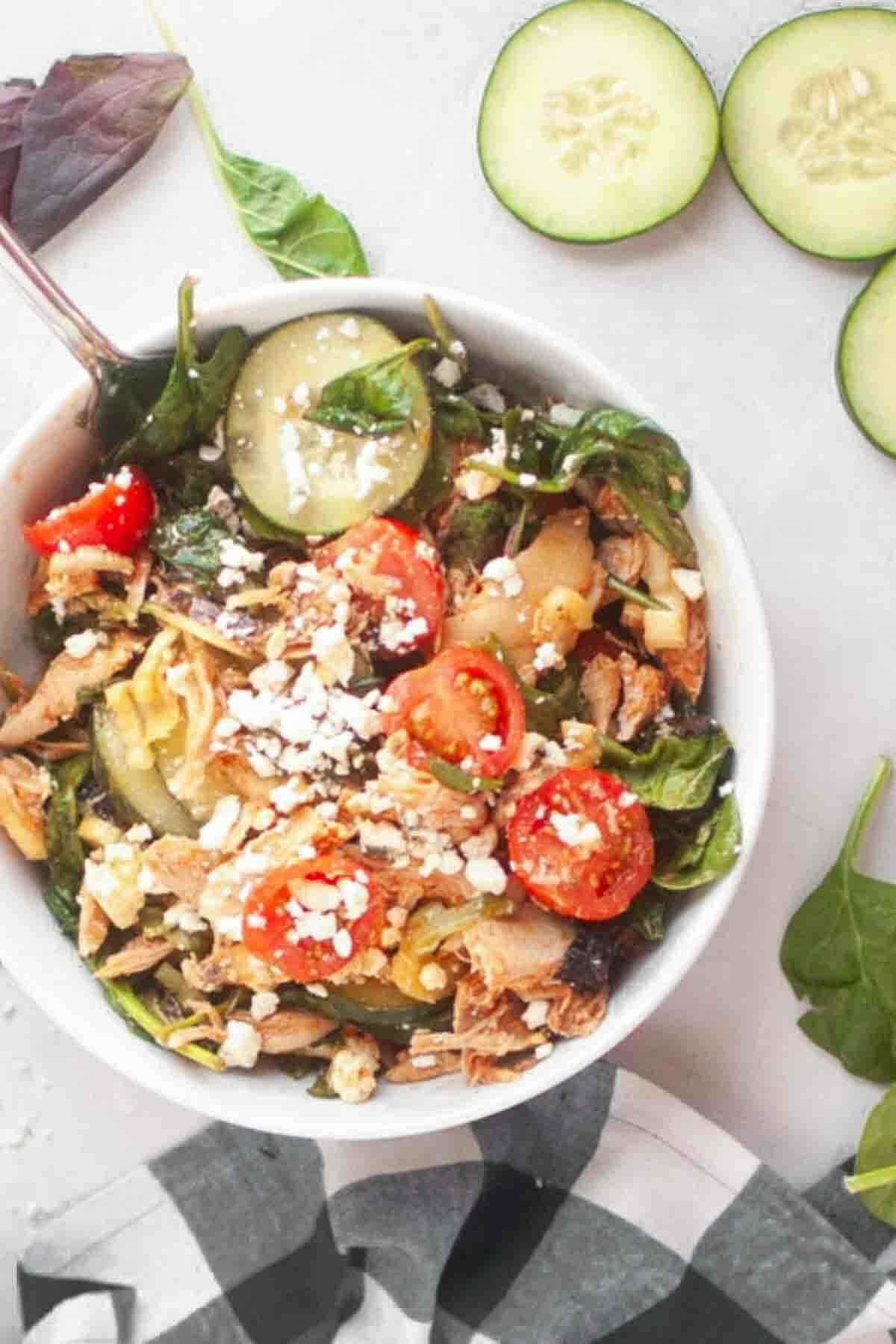 A bowl of salad with shredded chicken, cherry tomatoes, cucumber slices, spinach, and crumbled feta cheese, next to cucumber slices and spinach leaves on a white surface.