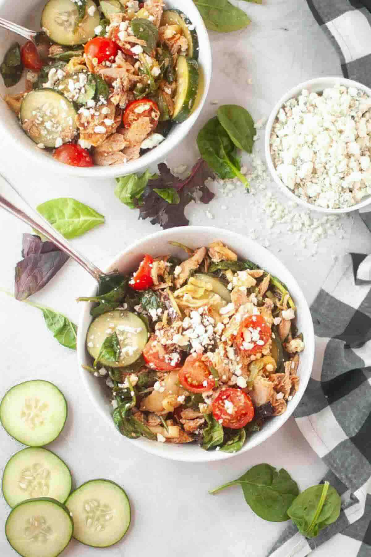 Two bowls of salad with sliced cucumber, cherry tomatoes, shredded chicken, greens, and crumbled feta cheese. A small bowl of feta and fresh cucumber slices are also visible.