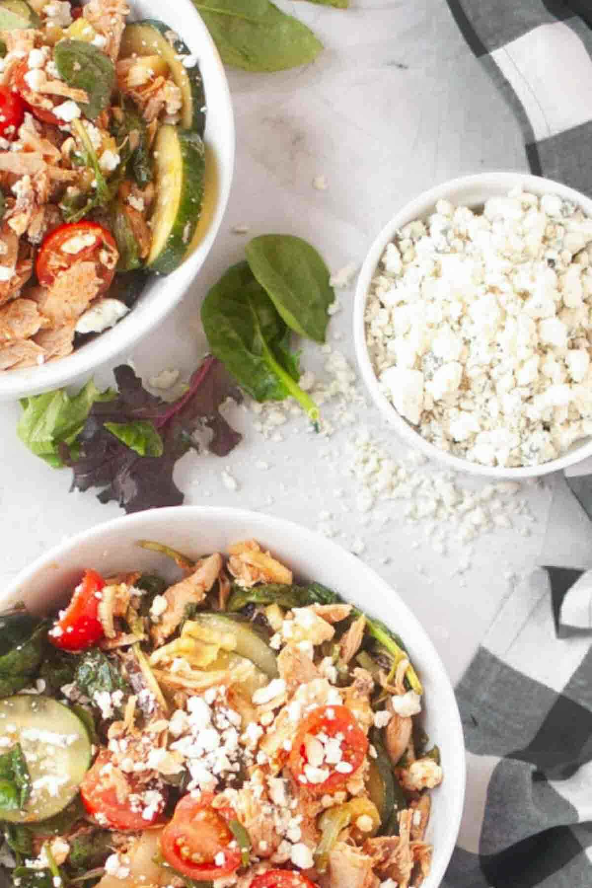 Two bowls of salad with shredded chicken, cherry tomatoes, zucchini, spinach, and crumbled cheese, next to a small bowl of extra crumbled cheese on a white surface.
