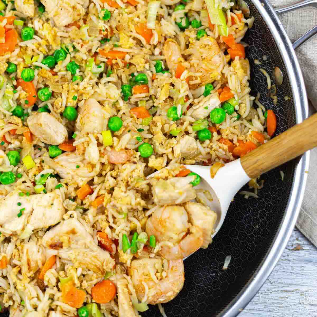 A skillet filled with fried rice, shrimp, chicken pieces, peas, carrots, and green onions, with a wooden-handled spoon scooping a portion.