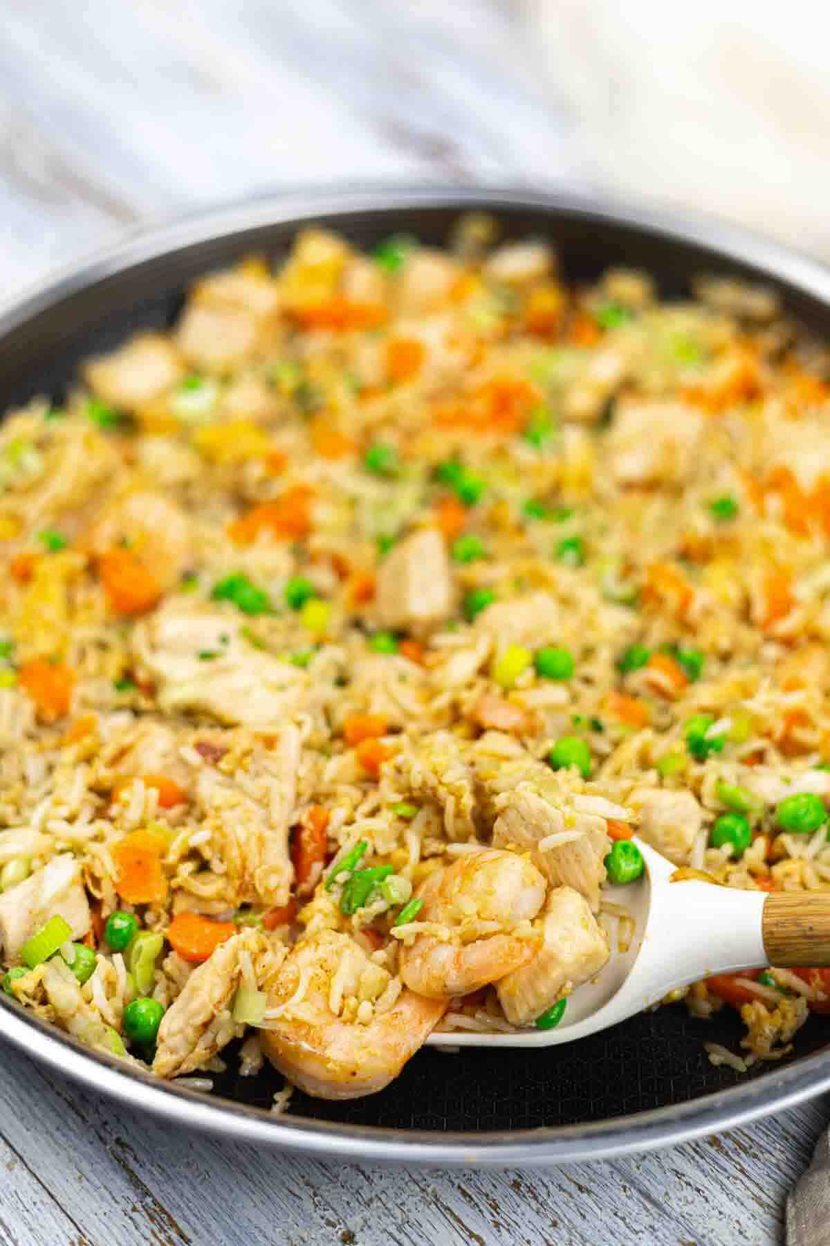 A pan filled with shrimp fried rice, including shrimp, chicken, peas, carrots, and rice; a spoon is lifting a portion from the pan.