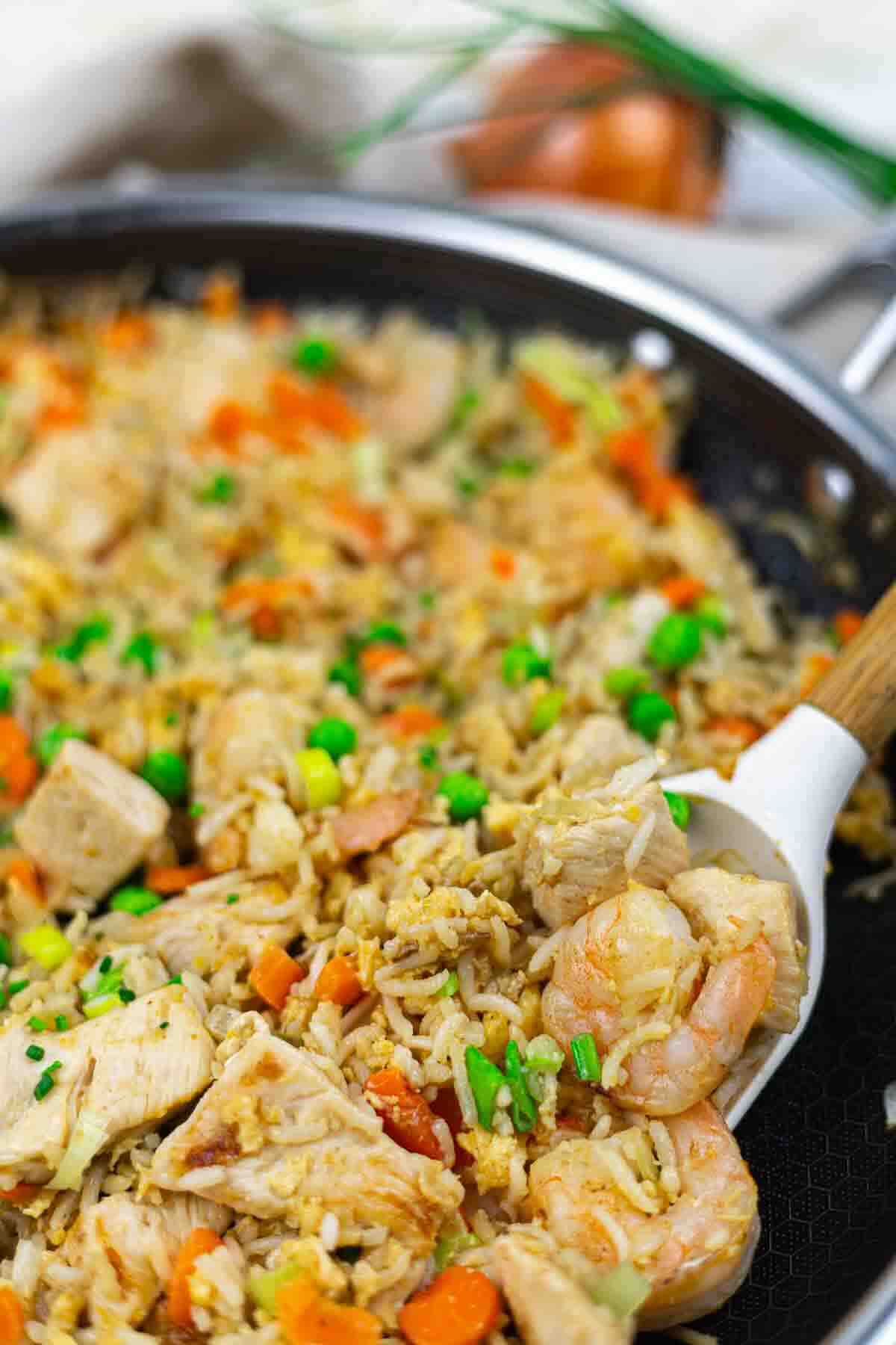 A skillet filled with fried rice, shrimp, diced chicken, carrots, peas, and green onions; a serving spoon is scooping some of the mixture.