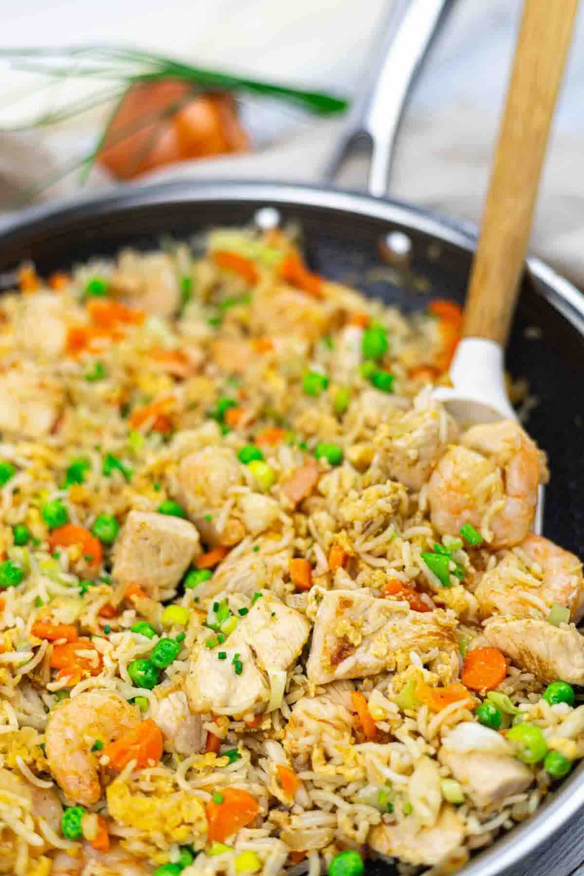 A skillet filled with fried rice, shrimp, chicken, peas, carrots, and scrambled eggs, with a wooden spoon resting inside.