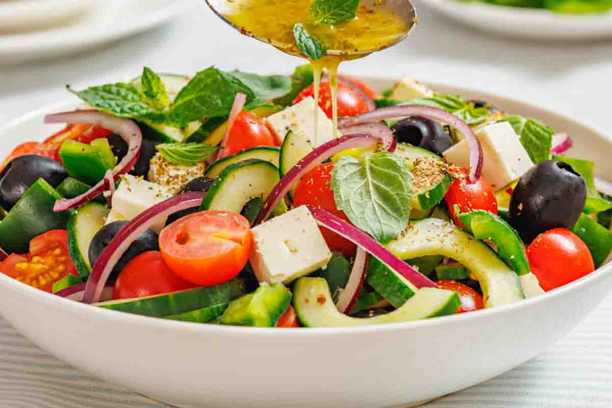 A bowl of Greek salad with tomatoes, cucumbers, red onions, olives, feta cheese, and mint leaves, being drizzled with dressing from a spoon.