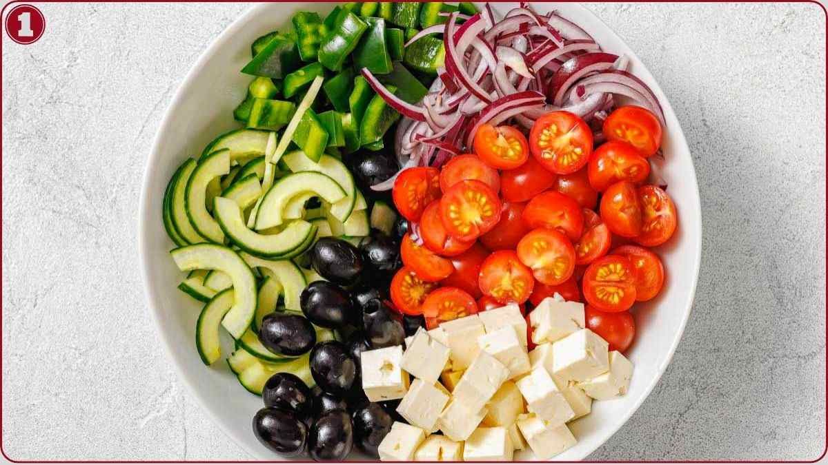 A bowl containing sliced cucumbers, green bell peppers, red onions, cherry tomatoes, black olives, and cubes of feta cheese arranged in sections.