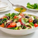 A spoon drizzles dressing over a bowl of Greek salad with tomatoes, cucumbers, feta cheese, olives, red onions, and fresh herbs.