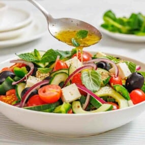 A spoon drizzles dressing over a bowl of Greek salad with tomatoes, cucumbers, feta cheese, olives, red onions, and fresh herbs.