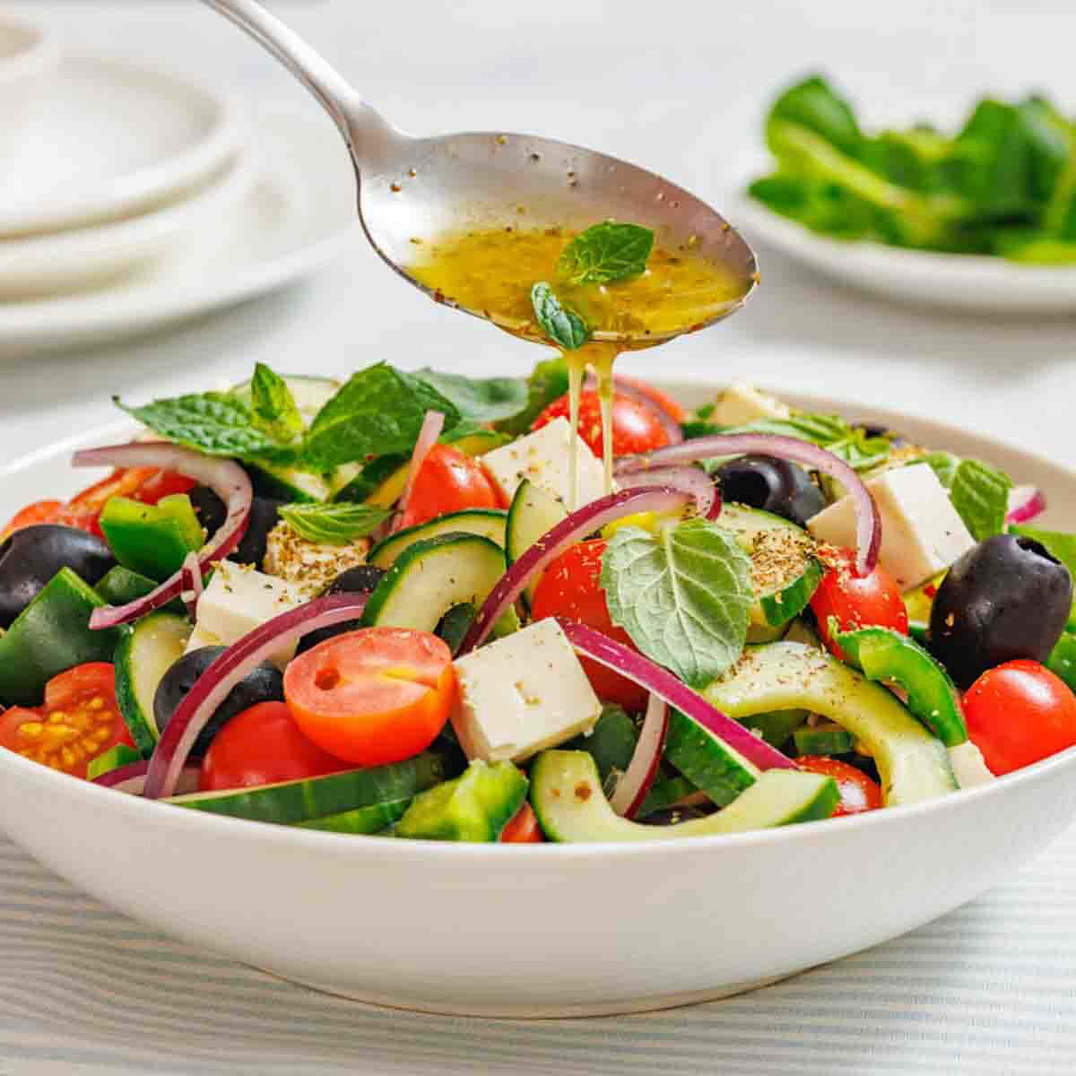 A spoon drizzles dressing over a bowl of Greek salad with tomatoes, cucumbers, feta cheese, olives, red onions, and fresh herbs.