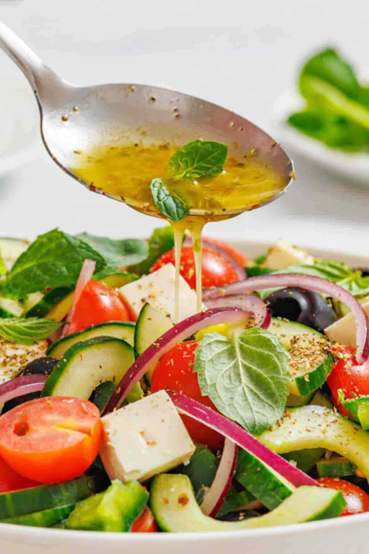 A spoon drizzles dressing over a bowl of Greek salad with tomatoes, cucumbers, red onions, olives, feta cheese, and fresh basil leaves.