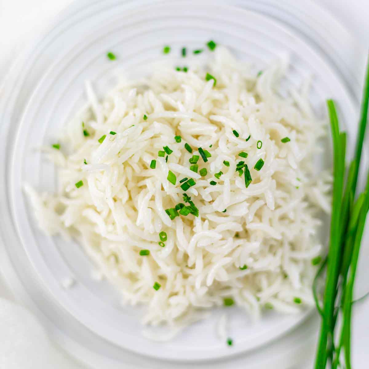A plate of cooked white rice garnished with chopped chives, with fresh chives placed beside the rice on a white plate.