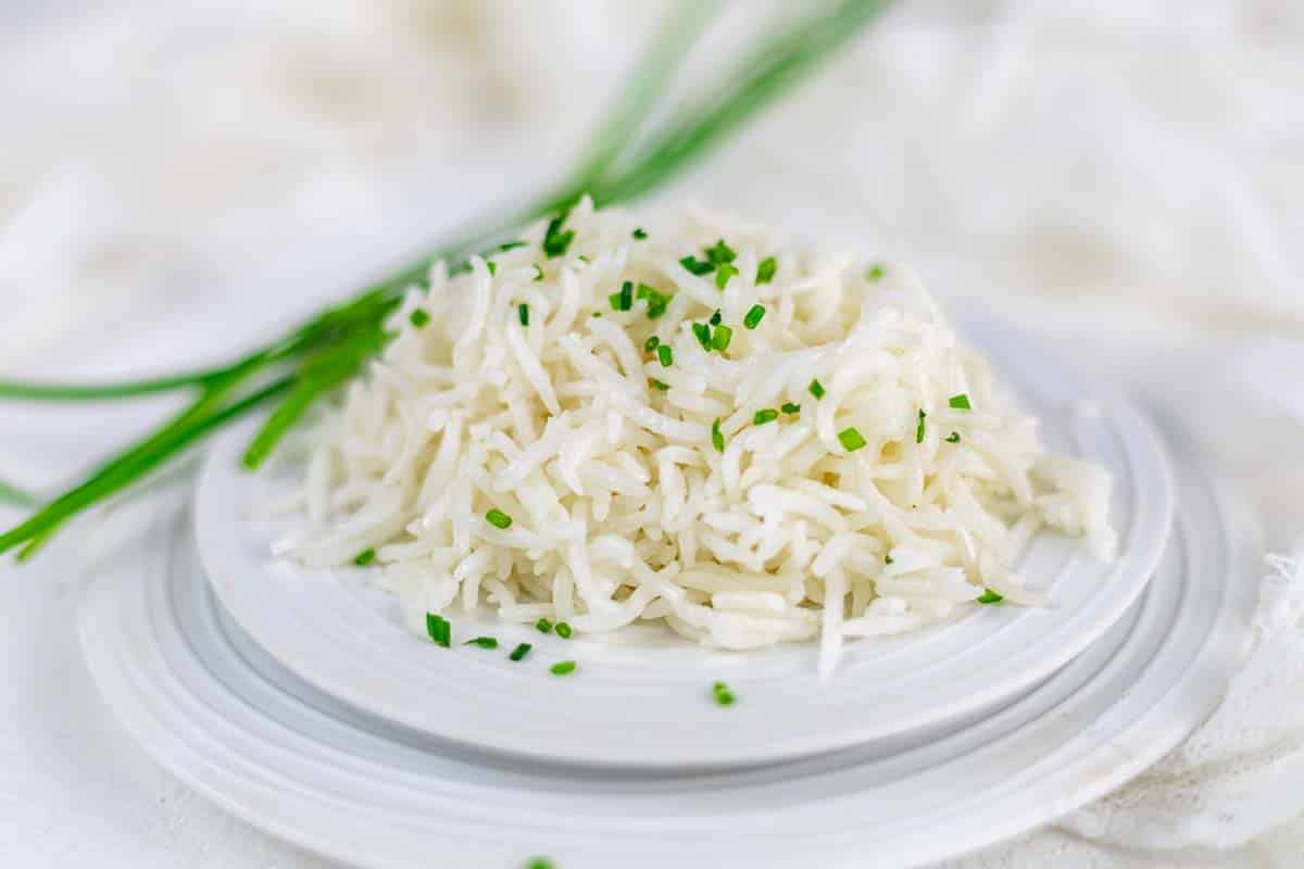 A serving of cooked white rice garnished with chopped chives on a white plate, with whole chives in the background.
