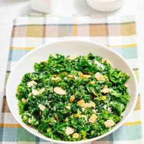 A white bowl filled with kale salad topped with sliced almonds and grated cheese, placed on a plaid cloth.